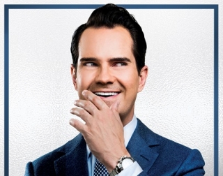 Jimmy Carr laughs into his hand whilst looking to the side, wearing a navy suit and a patterned tie. Jimmy Carr laughs into his hand whilst looking to the side, wearing a navy suit and a patterned tie.