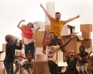 A man, woman and a group of children jump excitedly in the air in front of a pile of cardboard boxes. A man, woman and a group of children jump excitedly in the air in front of a pile of cardboard boxes.