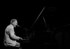 A man sings passionately into a mic on a piano. The photo is in black and white A man sings passionately into a mic on a piano. The photo is in black and white