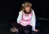 A female performer wearing a fluffy hat, white waterproof jacket and walking boots sits on a black stage A female performer wearing a fluffy hat, white waterproof jacket and walking boots sits on a black stage