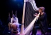 A woman plays the harp, a man smiles at her from behind an array of instruments on the stage A woman plays the harp, a man smiles at her from behind an array of instruments on the stage