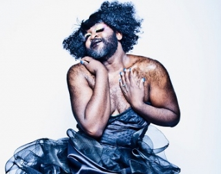 La Gateau Chocolat, a bearded black man with glitter in his hair, in a low cut black dress La Gateau Chocolat, a bearded black man with glitter in his hair, in a low cut black dress