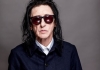 Dr John Cooper Clarke - a man with long hair and dark glasses - wears a dark blue and white shirt against a grey background. He is looking at the camera. Dr John Cooper Clarke - a man with long hair and dark glasses - wears a dark blue and white shirt against a grey background. He is looking at the camera.