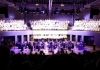 An orchestra on stage, with a choir dressed in white above them. An orchestra on stage, with a choir dressed in white above them.