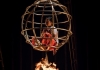 A child in a circular cage hanging above a fire installation and a woman dancing around it expressively, on a bed of exotic flowers A child in a circular cage hanging above a fire installation and a woman dancing around it expressively, on a bed of exotic flowers