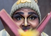 A man with a bobble hat, fake eyelashes and golden eye-shadow blowing yellow feathers from a book looking at the viewer directly A man with a bobble hat, fake eyelashes and golden eye-shadow blowing yellow feathers from a book looking at the viewer directly