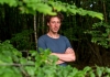 Sam Lee looking at the camera with his arms crossed in a foliage of woods Sam Lee looking at the camera with his arms crossed in a foliage of woods