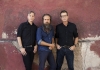 Three men in shirts and jeans lean against a red wall and stare at the camera Three men in shirts and jeans lean against a red wall and stare at the camera