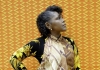 A woman in a a patterned shirt stands against a patterned yellow background A woman in a a patterned shirt stands against a patterned yellow background