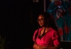 A woman wearing glasses and a red shirt stands on stage in front of a colourful backdrop A woman wearing glasses and a red shirt stands on stage in front of a colourful backdrop