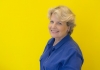 A woman wearing a blue shirt and standing in front of a yellow background smiles at the camera A woman wearing a blue shirt and standing in front of a yellow background smiles at the camera