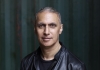 A man in a black t shirt and black jacket looks at the camera and stands in front of a striped green background A man in a black t shirt and black jacket looks at the camera and stands in front of a striped green background