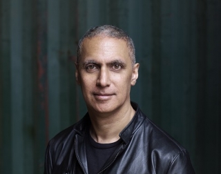 A man in a black t shirt and black jacket looks at the camera and stands in front of a striped green background A man in a black t shirt and black jacket looks at the camera and stands in front of a striped green background