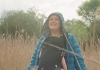 Josie Long is in a field of wheat smiling with her hood up and carrying a baby Josie Long is in a field of wheat smiling with her hood up and carrying a baby