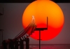 A man in a long gold robe climbs some stairs to a platform, with a large, red and orange sun in the background A man in a long gold robe climbs some stairs to a platform, with a large, red and orange sun in the background