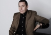 A man in dark jacket looks into camera whilst leaning on piano A man in dark jacket looks into camera whilst leaning on piano