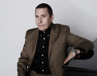 A man in dark jacket looks into camera whilst leaning on piano A man in dark jacket looks into camera whilst leaning on piano