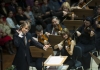 A conductor wearing a dark suit leads an orchestra. The strings section can be seen, with musicians playing violins A conductor wearing a dark suit leads an orchestra. The strings section can be seen, with musicians playing violins