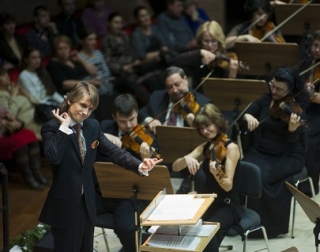 A conductor wearing a dark suit leads an orchestra. The strings section can be seen, with musicians playing violins A conductor wearing a dark suit leads an orchestra. The strings section can be seen, with musicians playing violins