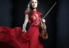 A woman with long brown hair, wearing a red dress stands against a black background, holding a violin A woman with long brown hair, wearing a red dress stands against a black background, holding a violin
