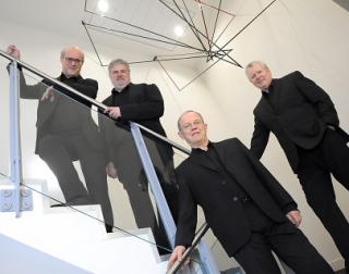 Four men in black suits stand on a glass staircase, underneath a metal sculpture, and look at the camera Four men in black suits stand on a glass staircase, underneath a metal sculpture, and look at the camera