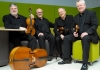 Four musicians in the black suits sit on bright green chairs with their instruments around them Four musicians in the black suits sit on bright green chairs with their instruments around them