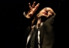 A performer stands on a black stage, wearing a black cape and white shirt, reaching upward A performer stands on a black stage, wearing a black cape and white shirt, reaching upward