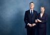A man and a woman dressed in dark suits stand arm in arm against a dark blue background. The man smiles at the camera as the woman looks away. A man and a woman dressed in dark suits stand arm in arm against a dark blue background. The man smiles at the camera as the woman looks away.