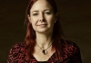 A woman with red hair smiles at the camera, standing in front of a black background A woman with red hair smiles at the camera, standing in front of a black background