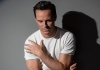 A man in a white t-shirt sat in front of a wall, looking down with his arm across his chest A man in a white t-shirt sat in front of a wall, looking down with his arm across his chest