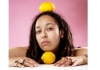 A person rests their chin on a lemon, looking at the camera, with another lemon resting on their head. A person rests their chin on a lemon, looking at the camera, with another lemon resting on their head.