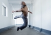 A performer jumps and twists in the air in a white and grey corridor. They are wearing grey trousers and a grey hood. A performer jumps and twists in the air in a white and grey corridor. They are wearing grey trousers and a grey hood.
