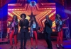 5 performers stand onstage and sing into microphones against a colourful background 5 performers stand onstage and sing into microphones against a colourful background