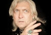 Clinton Baptiste against a black background, hand placed on his shoulder, looking past the camera Clinton Baptiste against a black background, hand placed on his shoulder, looking past the camera