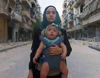 A photo of a woman carrying her baby set against the rubble and ruins of Aleppo A photo of a woman carrying her baby set against the rubble and ruins of Aleppo