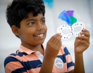 A smiling child holds an eye mask that has been decorated with gems and multi-coloured feathers A smiling child holds an eye mask that has been decorated with gems and multi-coloured feathers