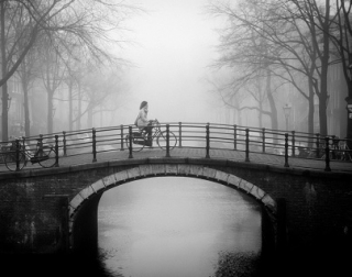 Black and white. A woman rides a bike over a canal bridge. It is foggy, and there are trees and buildings in the background. Black and white. A woman rides a bike over a canal bridge. It is foggy, and there are trees and buildings in the background.