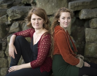 Two women sit back to back in front of a wall looking at the camera Two women sit back to back in front of a wall looking at the camera