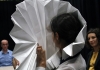 A woman with dark hair holds a large white paper fan in front of her face, watched by audience members. A woman with dark hair holds a large white paper fan in front of her face, watched by audience members.