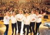 Six men stand on a stage looking at the camera, wearing white shirts and black trousers. There are instruments in the foreground and an audience in the background. Six men stand on a stage looking at the camera, wearing white shirts and black trousers. There are instruments in the foreground and an audience in the background.