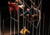 4 performers crouch on poles of scaffolding, high above a performer below 4 performers crouch on poles of scaffolding, high above a performer below