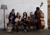 A group of musicians sit on a sofa, holding instruments such as a banjo, guitars and a bass as they look at each other and smile. A group of musicians sit on a sofa, holding instruments such as a banjo, guitars and a bass as they look at each other and smile.