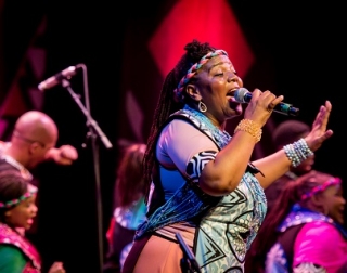 A woman in bright colourful dress sings into a microphone, with singers and dancers in the background A woman in bright colourful dress sings into a microphone, with singers and dancers in the background
