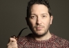 A person with short dark hair, wearing a knitted brown jumper, looks at the camera and holds a pipe A person with short dark hair, wearing a knitted brown jumper, looks at the camera and holds a pipe