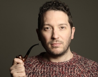 A person with short dark hair, wearing a knitted brown jumper, looks at the camera and holds a pipe A person with short dark hair, wearing a knitted brown jumper, looks at the camera and holds a pipe