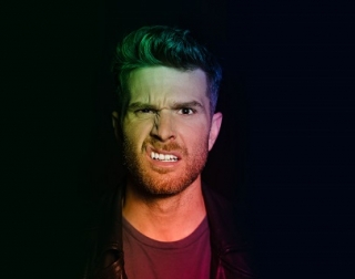 A man in a leather jacket stands against a black background, pulling a confused face at the camera. A man in a leather jacket stands against a black background, pulling a confused face at the camera.