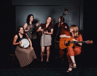 A group of women hold their instruments and laugh and talk together, in a dark room with a patterned carpet. A group of women hold their instruments and laugh and talk together, in a dark room with a patterned carpet.