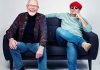 A photo of two men, sat on a sofa smiling, one of them is wearing a red hat and round sunglasses A photo of two men, sat on a sofa smiling, one of them is wearing a red hat and round sunglasses