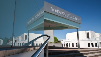 An image showing Warwick Arts Centre from the outside. A set of steps is pictured, with a handrail going up, and a grey sign that reads "Warwick Arts Centre". An image showing Warwick Arts Centre from the outside. A set of steps is pictured, with a handrail going up, and a grey sign that reads "Warwick Arts Centre".