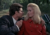 A man in a suit and a woman with blonde hair wearing a red coat (Deneuve) sit in the back of an open top car. A man in a suit and a woman with blonde hair wearing a red coat (Deneuve) sit in the back of an open top car.